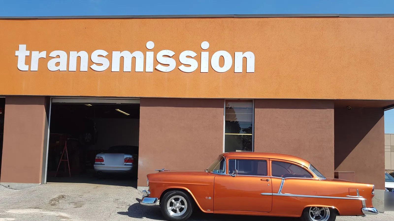 The front of a repair shop with a large orange wall, an open service bay with a car inside, and a restored orange classic car parked outside