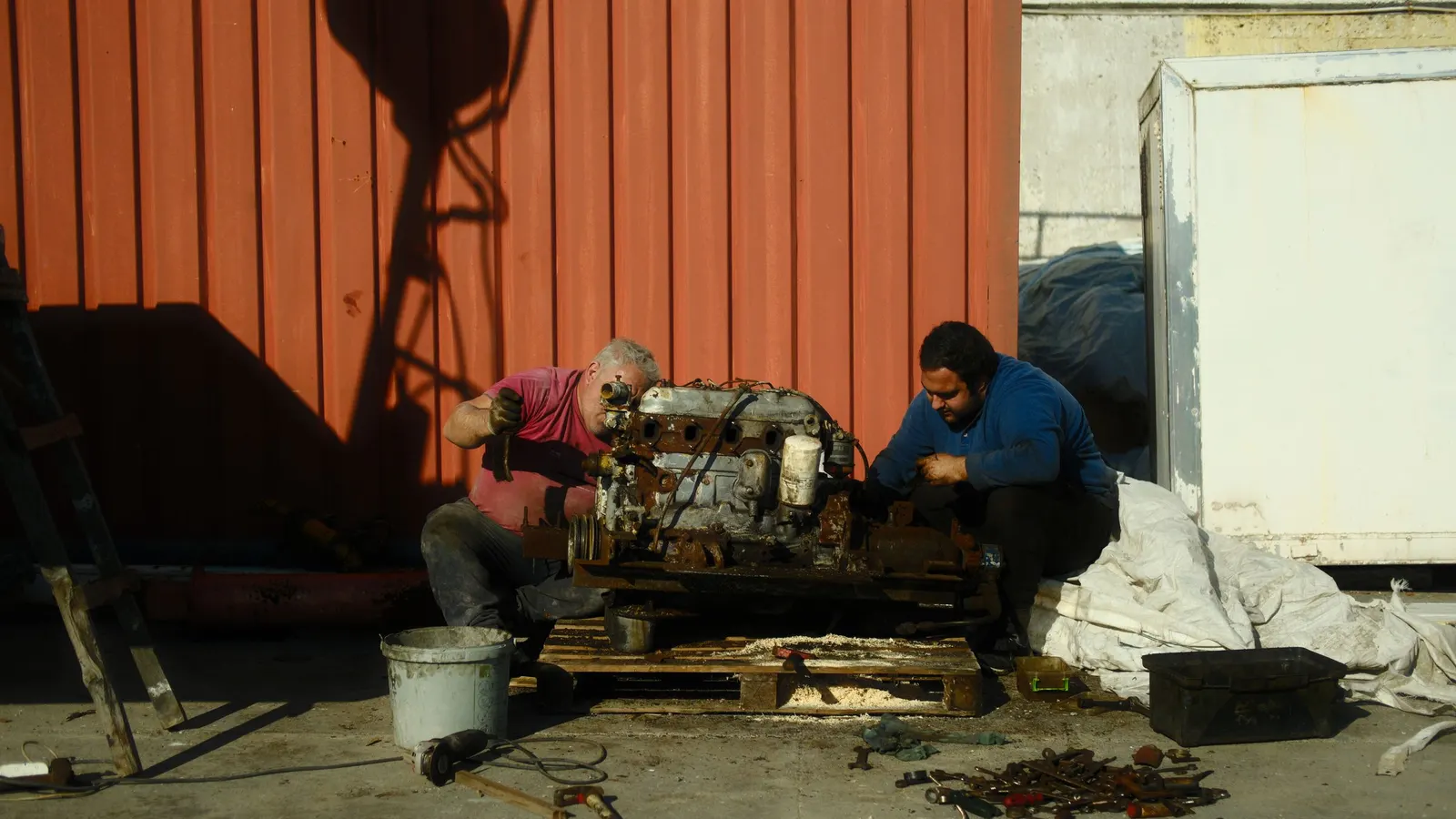 Two mechanics crouched outdoors, working together on an old engine set on a wooden pallet, with tools spread out on the ground